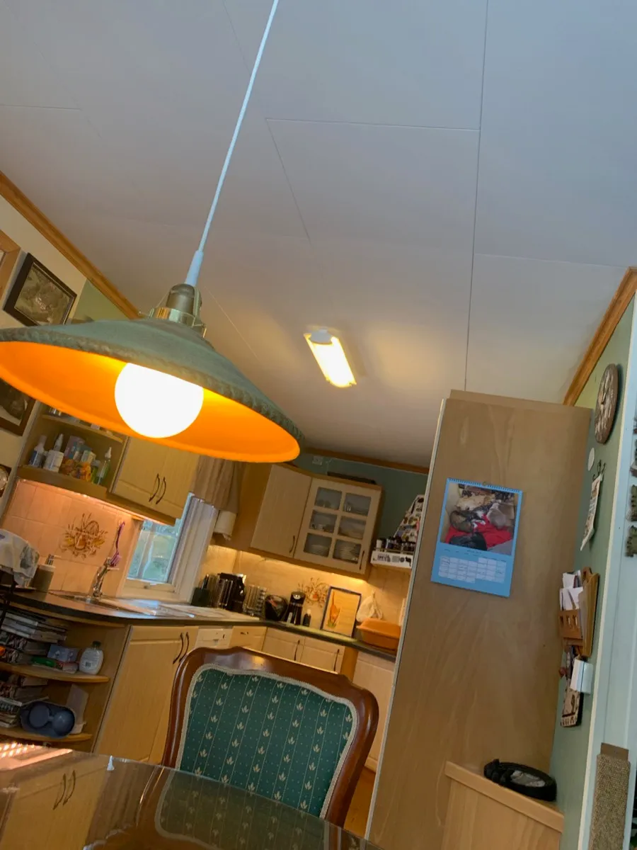Kjøkkenet sett fra spisebordet, med en taklampe med grønn skjerm og en blank lyspære i forgrunnen og en takarmatur med lysrør over kjøkkenbenken