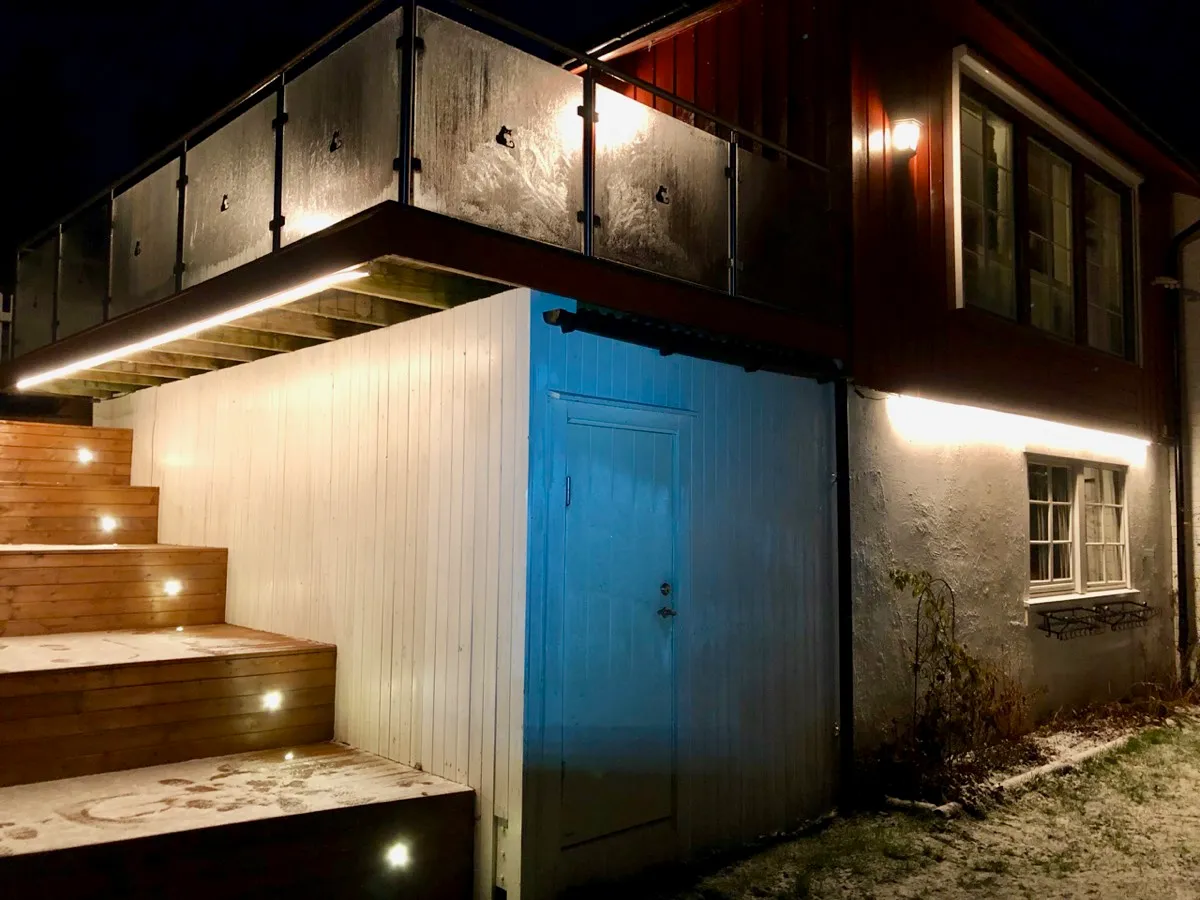 Husvegg om kvelden med lysstriper under verandaen og langs grunnmuren, og trapp med innfelte trinnlys