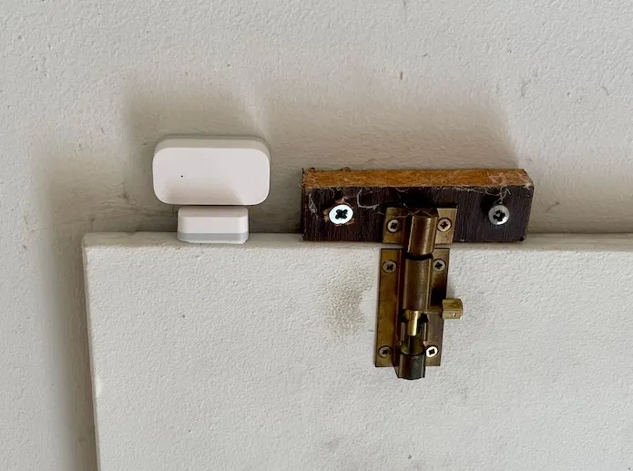 Liten hvit Aqara kontaktsensor montert på kanten av loftsluken, ved siden av en messingskåte