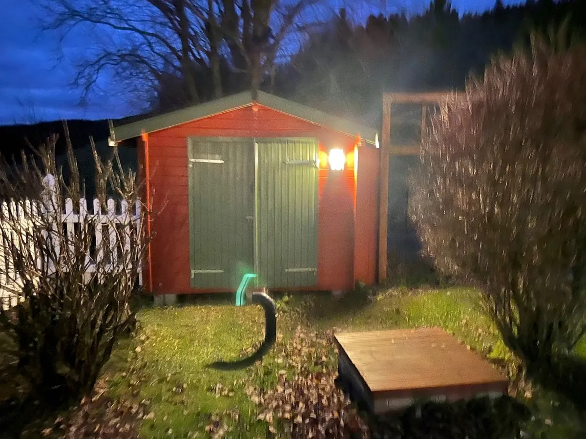 Rød bod med grønne dører og tent vegglampe, sett fra hagen i skumringen