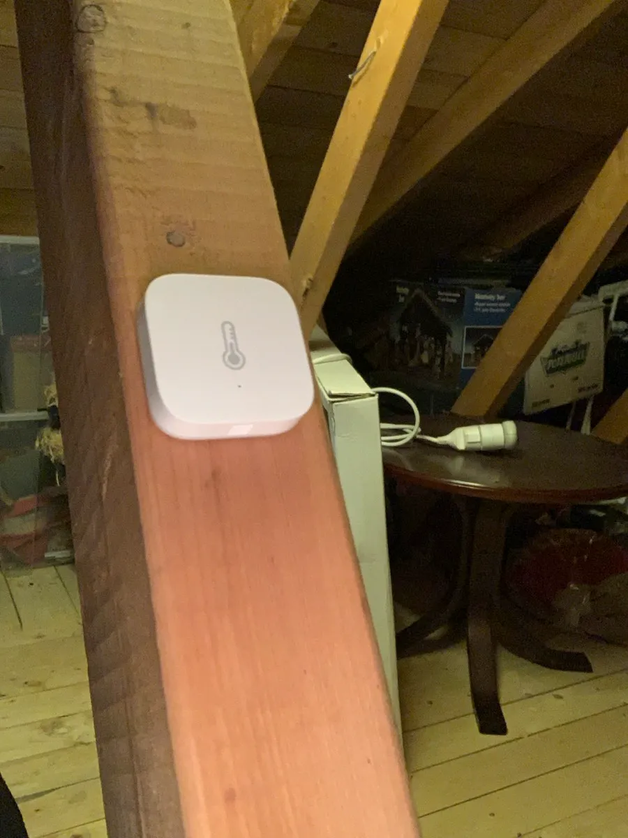 Liten, kvadratisk hvit Aqara-sensor med termometersymbol, festet på en trebjelke på loftet