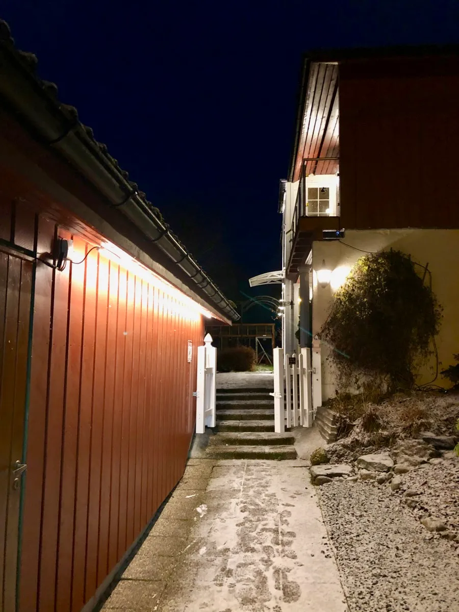 Smal passasje mellom garasjen og huset om kvelden, opplyst av en lysstripe under takskjegget på garasjen