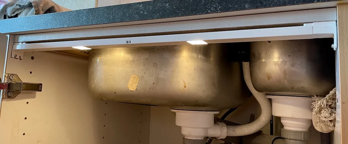 Skap under kjøkkenvasken, med to små lyspunkter i en hvit list under benkeplaten