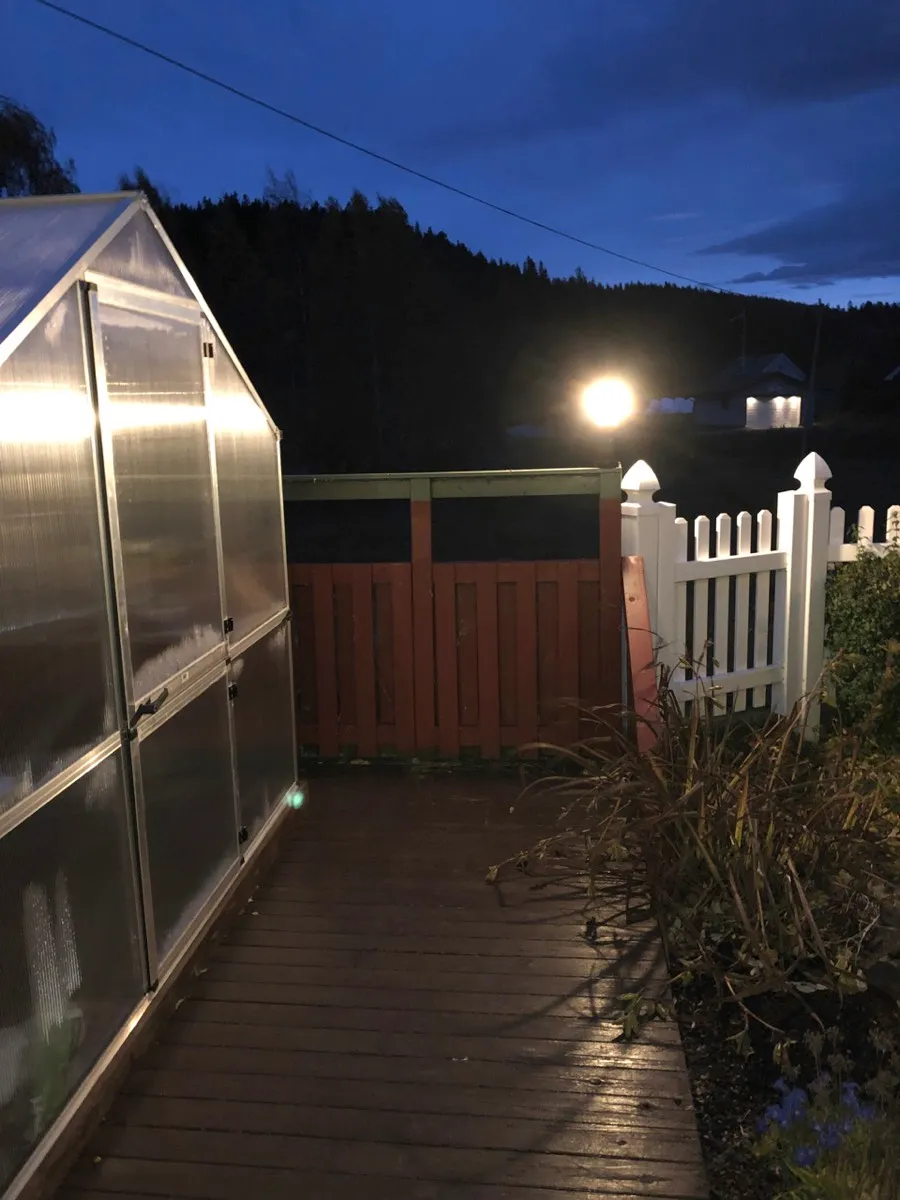 Drivhus og terrasse i skumringen, med en tent lyktestolpe ved hageporten